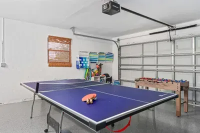 Spacious games room featuring a blue ping pong table at the center, ready for play with paddles and a ball resting on it. Adjacent to the table, a foosball table adds to the fun atmosphere.