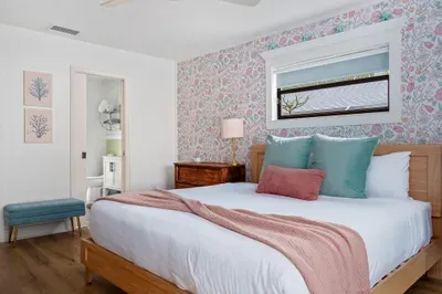 This image presents a comfortable bedroom featuring a large bed with crisp white sheets and soft pastel throws. The floral wallpaper continues to create warmth and charm, while a bedside table with a lamp provides ambient lighting.