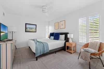 This photo features a spacious and well-lit bedroom, showcasing a neatly made bed and a television on a light wooden dresser. The calming blue and white color palette is complemented by nature-inspired art.