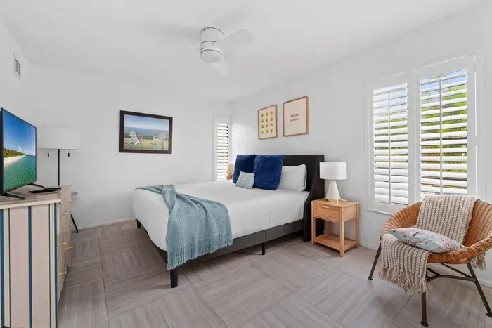 This photo features a spacious and well-lit bedroom, showcasing a neatly made bed and a television on a light wooden dresser. The calming blue and white color palette is complemented by nature-inspired art.
