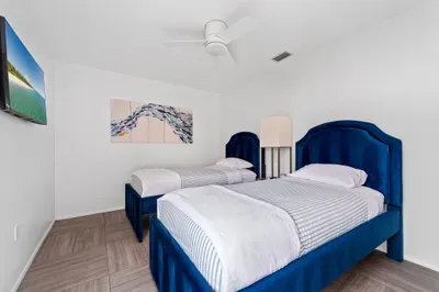 An overview of the bedroom, featuring the twin beds with plush bedding and a sleek bedside lamp. The bright white walls and wooden flooring create a clean look, while the artwork enhances the room's beach-themed decor.
