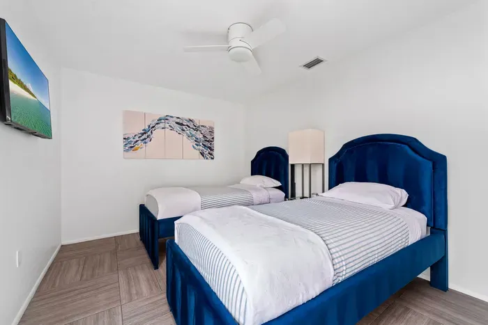 An overview of the bedroom, featuring the twin beds with plush bedding and a sleek bedside lamp. The bright white walls and wooden flooring create a clean look, while the artwork enhances the room's beach-themed decor.