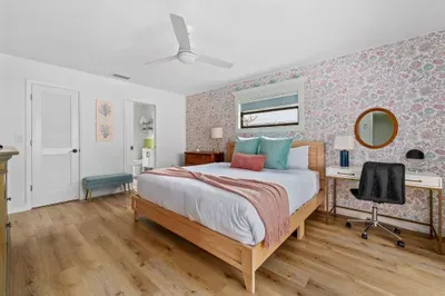 This image captures a well-lit bedroom featuring a stylish bed with assorted pillows. The floral wallpaper establishes a cheerful ambiance, complemented by a small desk and round mirror.
