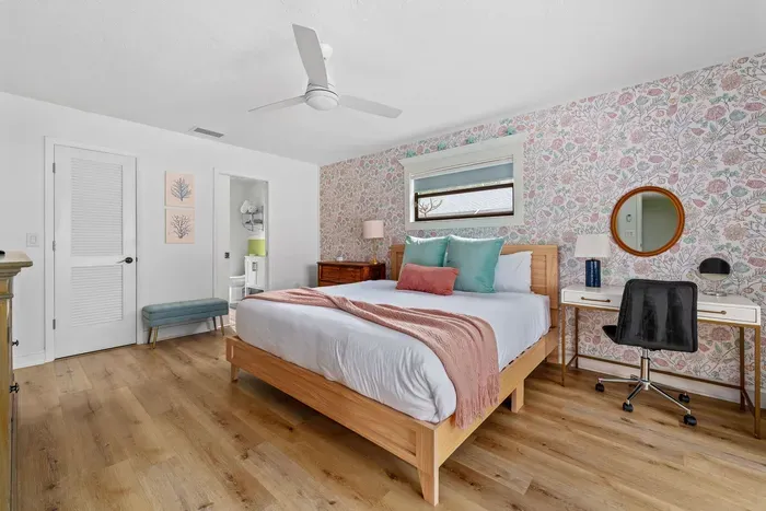 This image captures a well-lit bedroom featuring a stylish bed with assorted pillows. The floral wallpaper establishes a cheerful ambiance, complemented by a small desk and round mirror.