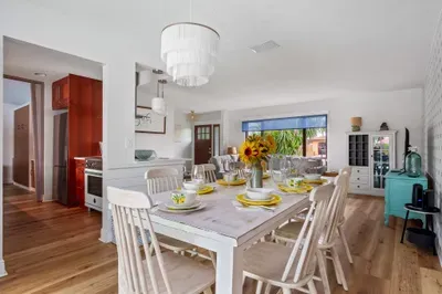 This image offers a wide view of the dining room, featuring a beautifully set table with colorful plates and clear glassware. The white walls and chandelier create a light, airy ambiance. A turquoise accent piece in the background adds a pop of color