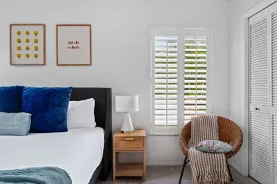 Features a bright bedroom corner with a large bed dressed in white linens and bold blue accent pillows. A stylish wooden nightstand holds a modern lamp. Nearby, a woven chair with a striped throw adds texture and comfort,