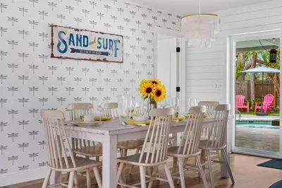 This photo highlights the dining room's cozy atmosphere, featuring the "Sand and Surf" sign and a table adorned with sunflowers. The open door leads to the backyard, where poolside seating can be seen, inviting relaxation.