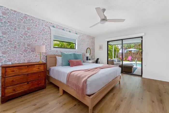 This photo shows a spacious bedroom filled with natural light. The floral wallpaper, soft bedding, and wooden dresser create a harmonious and cozy space. Large glass doors lead to an outdoor area, inviting guests to enjoy the fresh air.