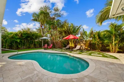 This photo showcases a tranquil outdoor space with two modern loungers positioned beside a sparkling swimming pool. A vibrant red umbrella provides shade, while lush greenery and tropical plants surround the area, creating a serene retreat.