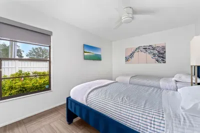 Captures a bright bedroom featuring two comfortable twin beds with crisp white linens and a subtle striped pattern. A ceiling fan hangs overhead, while a window offers a view of lush greenery outside