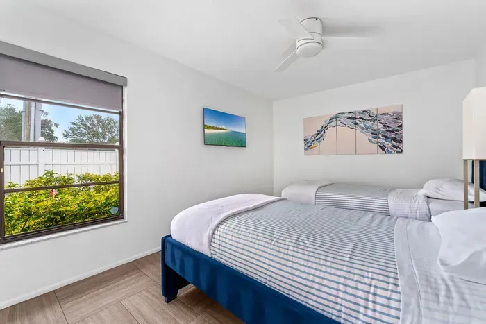 Captures a bright bedroom featuring two comfortable twin beds with crisp white linens and a subtle striped pattern. A ceiling fan hangs overhead, while a window offers a view of lush greenery outside