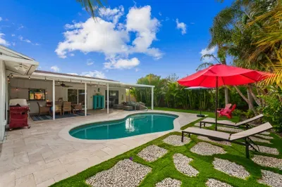 This inviting shot displays a panoramic view of the pool area, showcasing the loungers, the red umbrella, and hints of an outdoor dining space under a covered patio. The sky is clear, enhancing the relaxing vibe of the area.