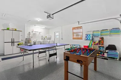 Spacious multi-purpose room featuring a ping pong table and a foosball table, perfect for indoor entertainment. The floor has a smooth finish, and a large refrigerator is visible in the background alongside other storage item