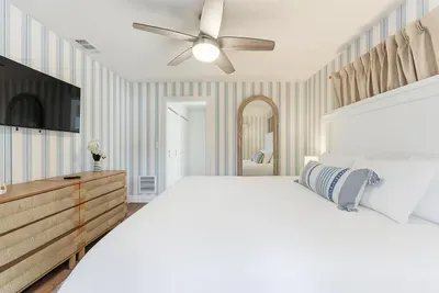 Coastal-inspired bedroom with striped walls, a plush king bed, and a stylish dresser with TV. Relax in this serene retreat that perfectly blends comfort, elegance, and beachy charm.