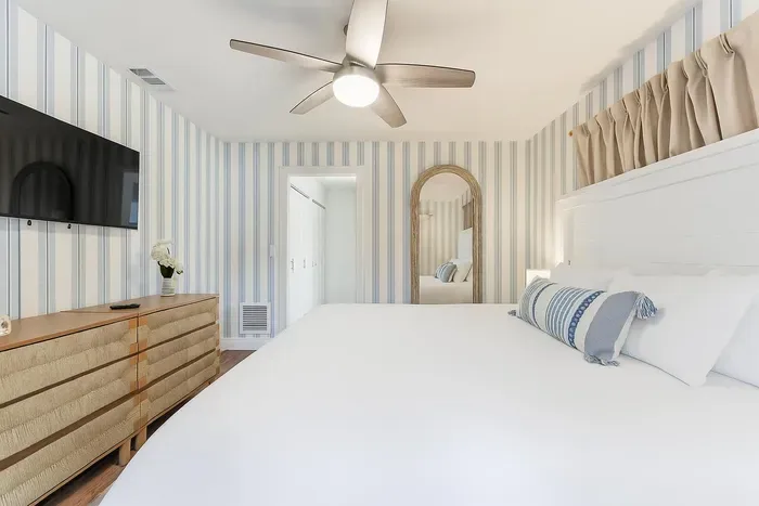 Coastal-inspired bedroom with striped walls, a plush king bed, and a stylish dresser with TV. Relax in this serene retreat that perfectly blends comfort, elegance, and beachy charm.