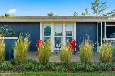 Charming curb appeal with modern blue tones, tall ornamental grasses, and a cozy patio accented by red Adirondack chairs. Perfect for morning coffee or evening relaxation, this welcoming front view sets the tone for a stylish coastal retreat.