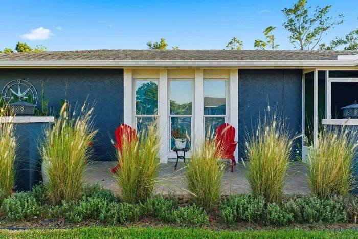 Charming curb appeal with modern blue tones, tall ornamental grasses, and a cozy patio accented by red Adirondack chairs. Perfect for morning coffee or evening relaxation, this welcoming front view sets the tone for a stylish coastal retreat.