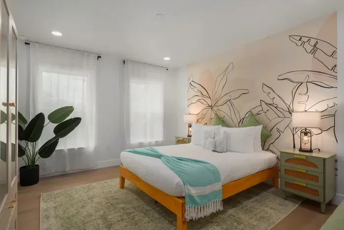 Tranquil bedroom retreat with plush bedding, soft lighting, and a beautiful tropical mural backdrop. Natural light, calming tones, and cozy textures create a peaceful space to unwind and recharge after beach-filled days.