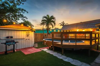 Magical backyard oasis at dusk with private pool, elevated deck, hot tub, BBQ grill, and cozy lounge seating. String lights, palm trees, and sunset skies set the scene for relaxing swims, evening cookouts, and unforgettable nights.