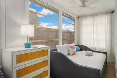 Sun-filled bedroom with cozy daybed-style seating, soft linens, and coastal accents. Large windows invite natural light, creating a cheerful, relaxing space perfect for lounging, reading, or peaceful sleep.