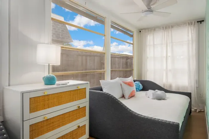Sun-filled bedroom with cozy daybed-style seating, soft linens, and coastal accents. Large windows invite natural light, creating a cheerful, relaxing space perfect for lounging, reading, or peaceful sleep.