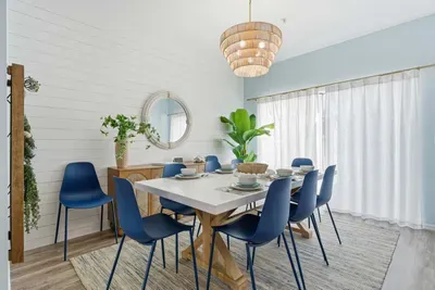 Bright coastal dining space with a modern farmhouse table, chic blue chairs, and natural textures. Sunlight pours through sheer curtains, creating the perfect setting for family meals, morning coffee, or relaxed gatherings in breezy beach style.