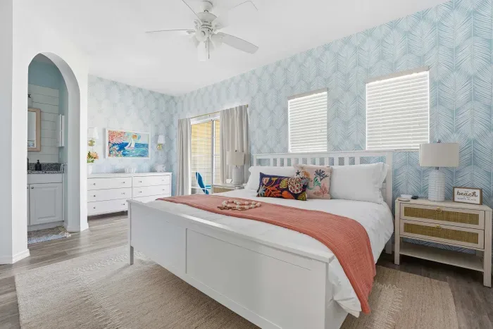 Serene coastal bedroom with a king bed, ocean-inspired wallpaper, and soft coral accents. Cozy lighting and beachy details create a peaceful space to unwind and drift off to the sound of nearby waves.