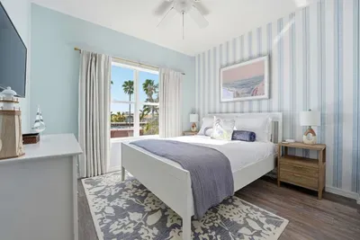 Soft blue tones, striped accent walls, and ocean-inspired artwork create a serene escape. Featuring plush bedding, warm wood nightstands, and abundant natural light, this relaxing retreat is perfect for peaceful mornings and restful nights.