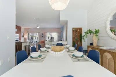 Bright and inviting dining area with a sleek white table, navy chairs, and coastal accents. Open to the living space, it’s perfect for family dinners, game nights, or gathering after a day at the beach.
