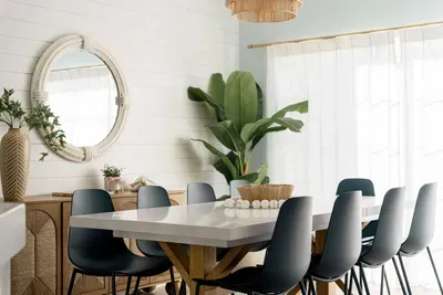 Featuring a sleek wood table, modern black chairs, statement mirror, and lush greenery. Bathed in natural light, this inviting setting blends comfort and design for relaxed breakfasts or elegant dinners.