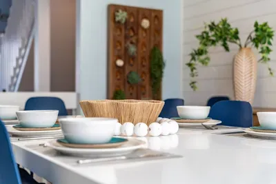 Elegant coastal dining setup with modern tableware, blue chairs, and natural accents. Thoughtful details and soft tones create a welcoming space perfect for shared meals, conversation, and relaxed seaside dining.