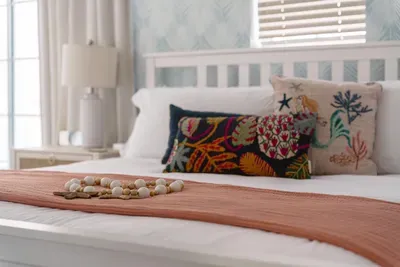 Crisp white linens, a soft coral throw, and vibrant seaside accent pillows create a warm, inviting feel. Thoughtfully styled with natural light and charming décor, this serene space is perfect for restful nights and slow mornings.