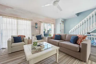 Bright, coastal-inspired living room filled with natural light. Featuring plush sofas with vibrant accent pillows, a stylish coffee table, and soft wood flooring, this inviting space is perfect for relaxing, gathering and unwinding after a sun-soaked