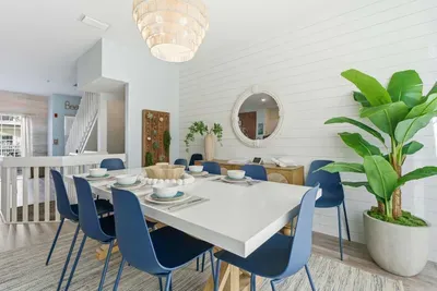 Modern coastal dining room featuring a sleek white table, navy chairs, and tropical greenery. Soft natural light and chic décor create the perfect space for family meals or relaxed gatherings by the beach.