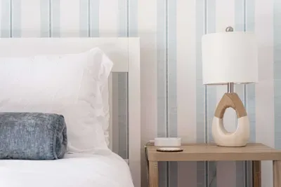 Crisp white linens pair beautifully with soft blue accents and serene striped walls. A warm wood nightstand and elegant lamp add charm and comfort, creating a calming, light-filled space perfect for restful nights and peaceful mornings.