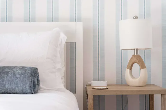 Crisp white linens pair beautifully with soft blue accents and serene striped walls. A warm wood nightstand and elegant lamp add charm and comfort, creating a calming, light-filled space perfect for restful nights and peaceful mornings.