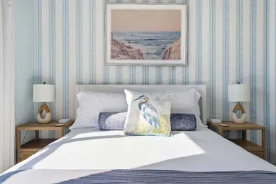 Framed by soft blue striped walls and ocean-inspired artwork, this serene retreat features plush bedding, elegant nightstands, and warm lighting. A peaceful haven designed for restful nights and refreshing mornings.