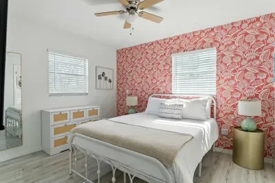 Stylish bedroom featuring a bold red floral accent wall, plush white bedding, and soft beige accents. Natural light, modern décor, and a ceiling fan create the perfect blend of comfort and charm for your coastal escape.