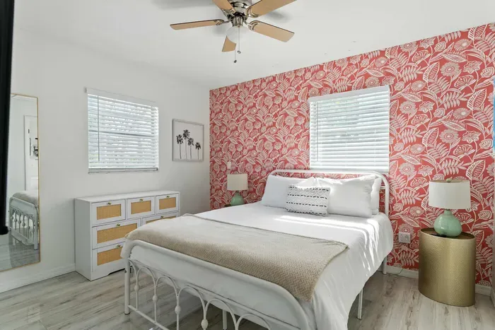 Stylish bedroom featuring a bold red floral accent wall, plush white bedding, and soft beige accents. Natural light, modern décor, and a ceiling fan create the perfect blend of comfort and charm for your coastal escape.