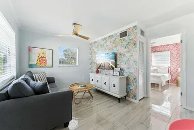 Stylish living space with a vibrant tropical accent wall, sleek tile floors, and a cozy gray sofa. Natural light flows in, leading to a charming bedroom—perfect for relaxing after sun-soaked beach adventures.