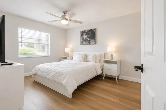 Bright and serene bedroom featuring a cozy queen bed, soft white linens, and warm wood flooring. Natural light pours through a large window, complemented by modern nightstands, soft lamps, and a calming neutral palette—perfect for restful nights.