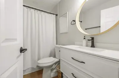 Stylish and spotless bathroom with a modern touch—featuring a sleek vanity, round illuminated mirror, and bright white finishes. Enjoy a refreshing shower behind the crisp curtain in this clean, inviting, spa-like space.