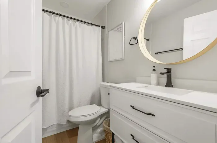 Stylish and spotless bathroom with a modern touch—featuring a sleek vanity, round illuminated mirror, and bright white finishes. Enjoy a refreshing shower behind the crisp curtain in this clean, inviting, spa-like space.