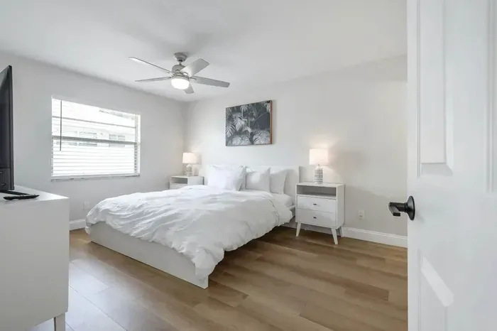 Bright and serene bedroom featuring a cozy queen bed, soft white linens, and warm wood flooring. Natural light pours through a large window, complemented by modern nightstands, soft lamps, and a calming neutral palette—perfect for restful nights.