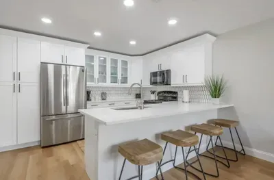 Bright and modern kitchen featuring sleek white cabinetry, stainless steel appliances, and a spacious breakfast bar with rustic stools. Perfect for cooking, dining, and entertaining in style.