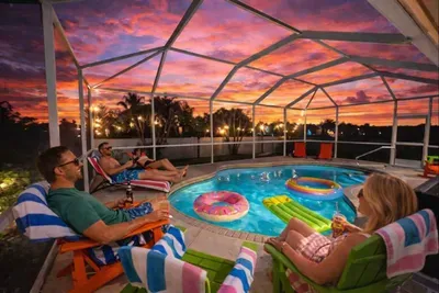 Screened-in pool oasis at sunset with glowing water, fun floats, and comfy Adirondack chairs. Guests unwind with drinks and towels, surrounded by palm views and warm lighting—perfect for relaxing evenings, family time, and memorable vacation vibes.