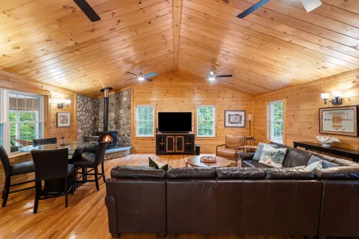 A warm, spacious living room with vaulted wood ceilings, big picture windows, cozy leather seating, and peaceful forest views. Enjoy the stone fireplace, wood-burning stove, and family-friendly lounge—perfect for movies, coffee, and relaxing together