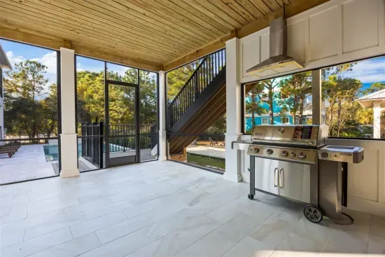 A breezy covered outdoor kitchen designed for effortless entertaining, featuring a premium gas grill beneath a wood-paneled ceiling and surrounded by tranquil greenery—perfect for relaxed coastal dinners just steps from the patio.
