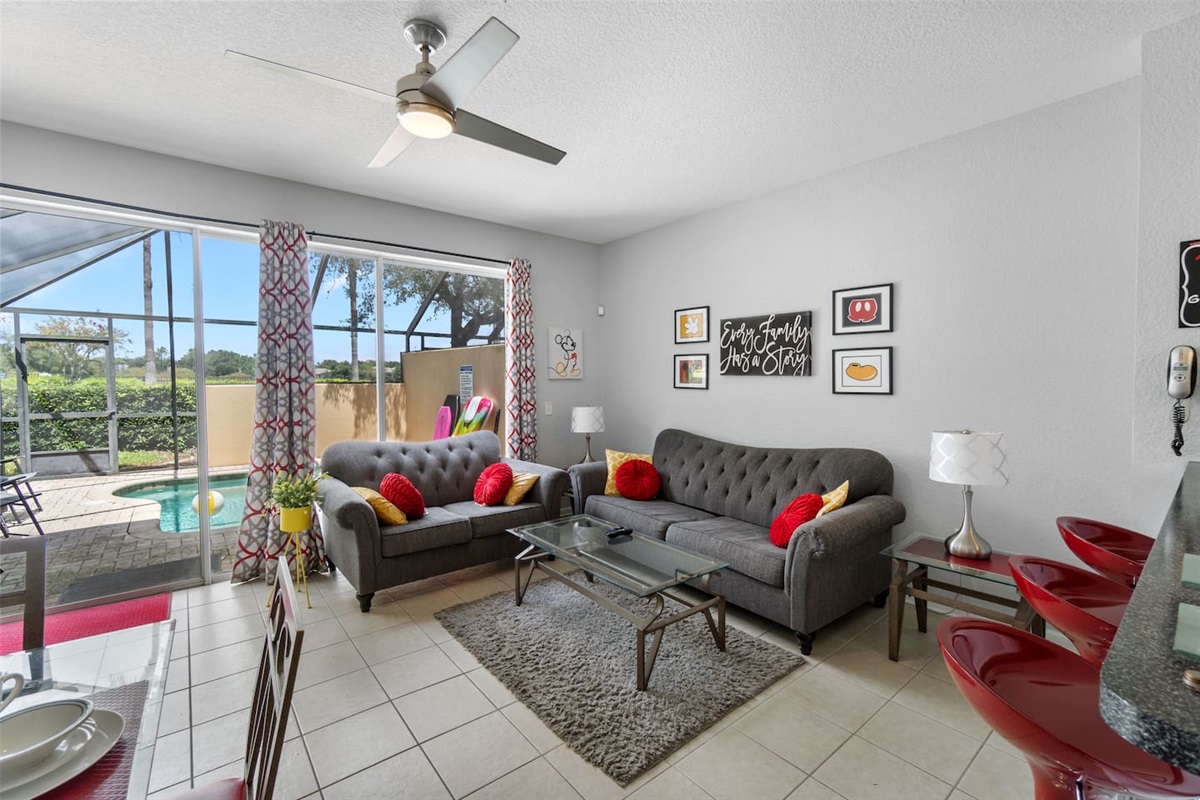 - Bright living room with plush gray sofas, vibrant red cushions, and wall art  
- Sliding doors open to the pool area for easy indoor outdoor flow and relaxation  
- Ceiling fan and natural light create a fresh, welcoming atmosphere year round