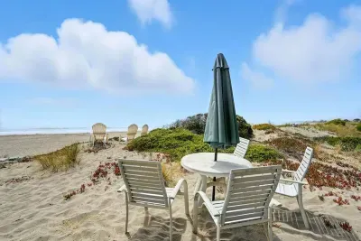 Outdoor dining set on sandy beach with umbrella shade.